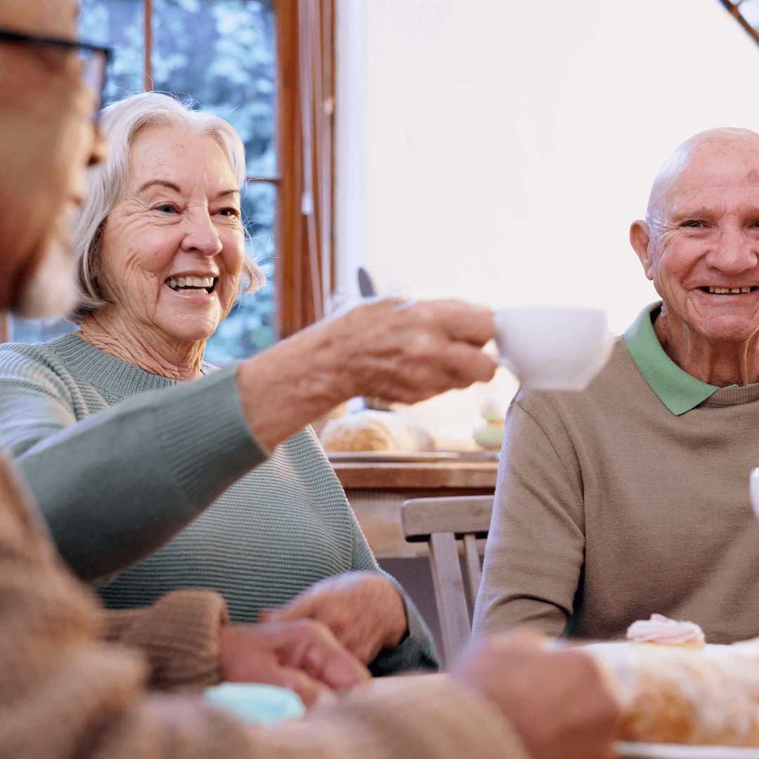 Two elderly people happily chat while sharing a cup of tea at a table in a cozy room with a window in the background. - Home Instead