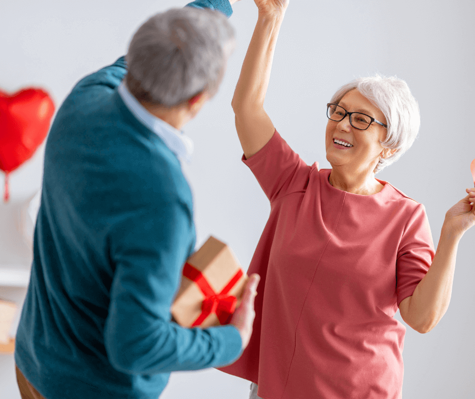 Elderly couple joyfully dancing; woman smiling, man holding a gift with a red ribbon. Red heart decoration in the background. - Home Instead