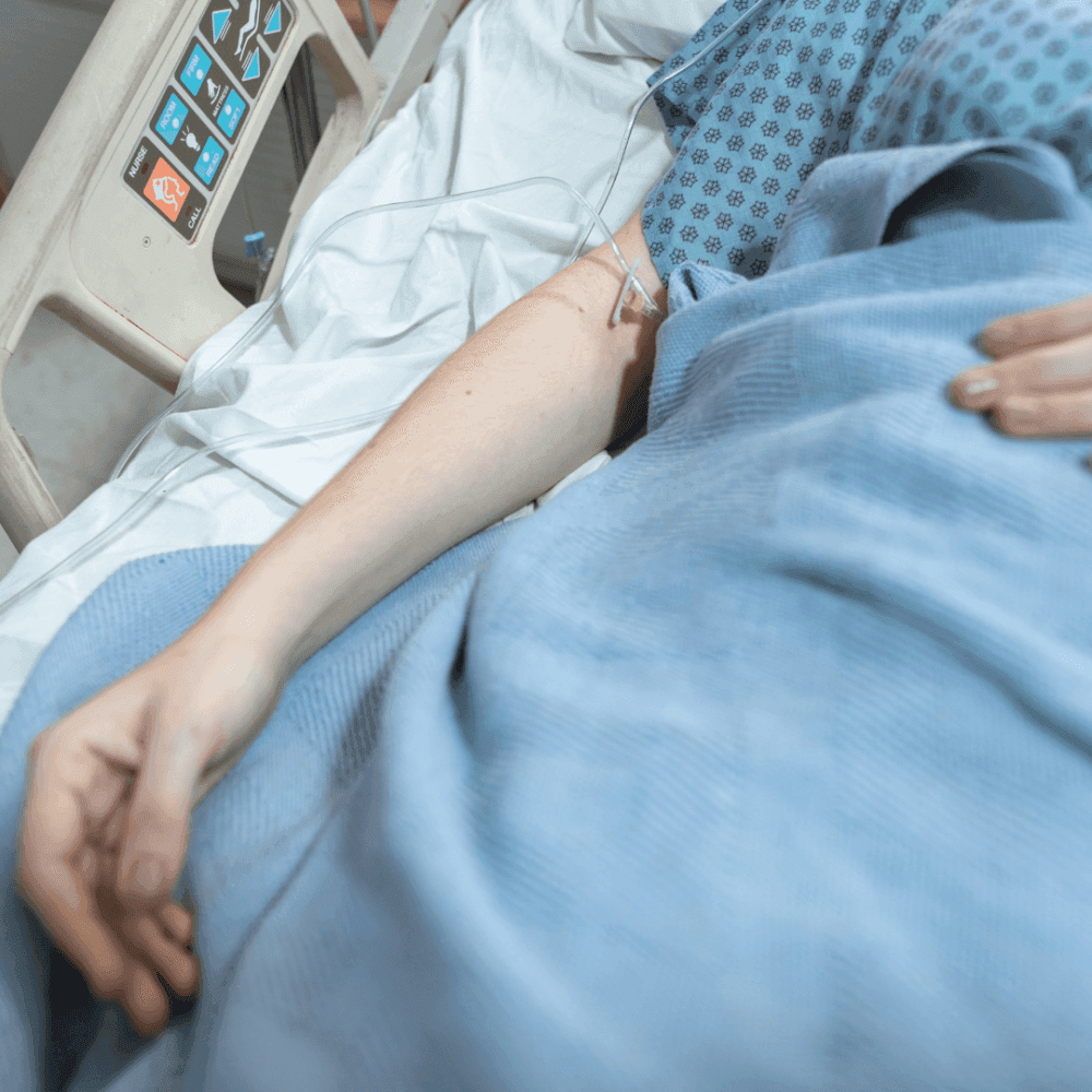 Person lying in a hospital bed with an arm resting outside the blanket, an IV is attached to their hand. - Home Instead