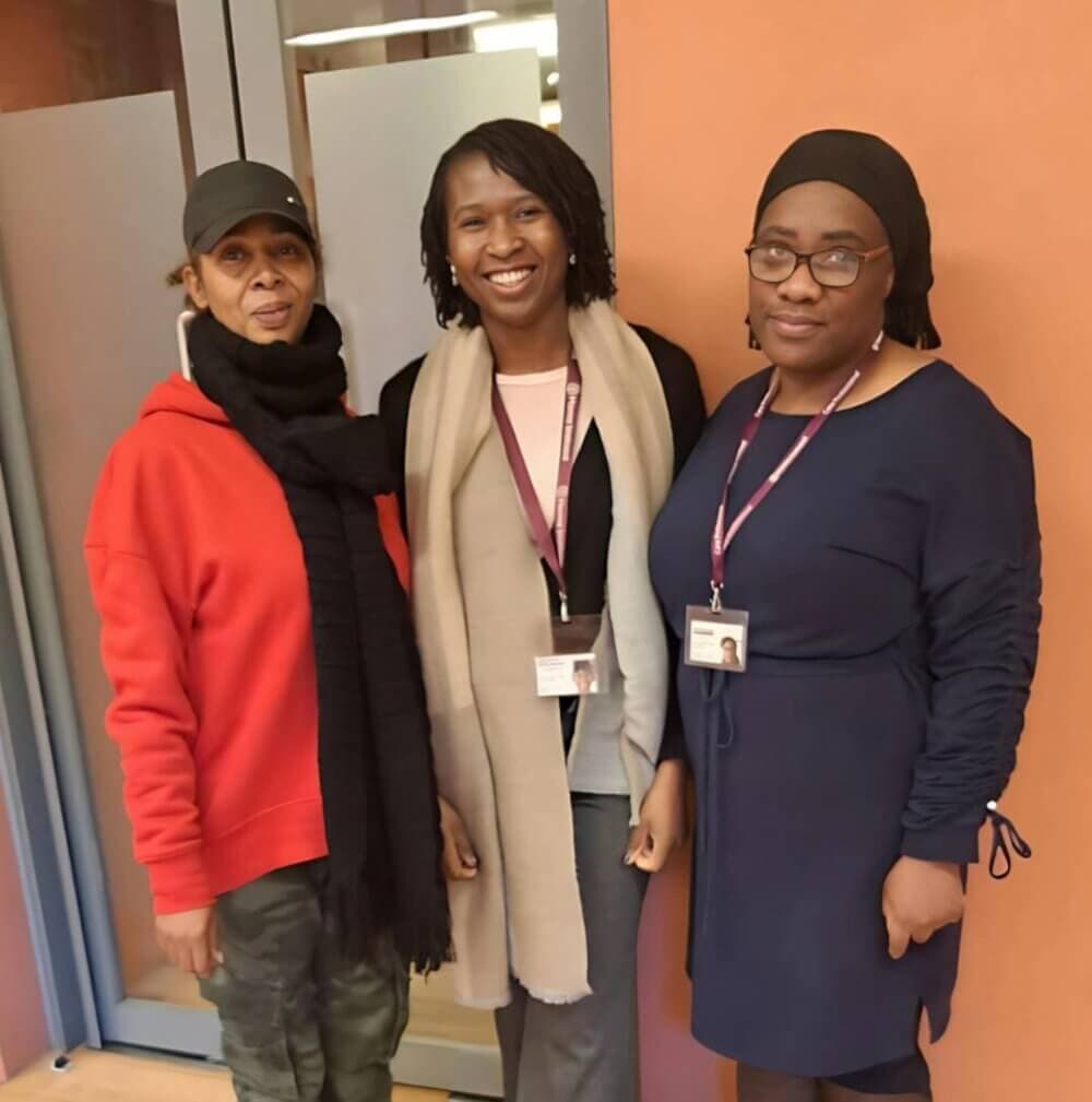 Three women standing together, smiling, wearing ID badges with lanyards, in front of a beige and orange background. - Home Instead