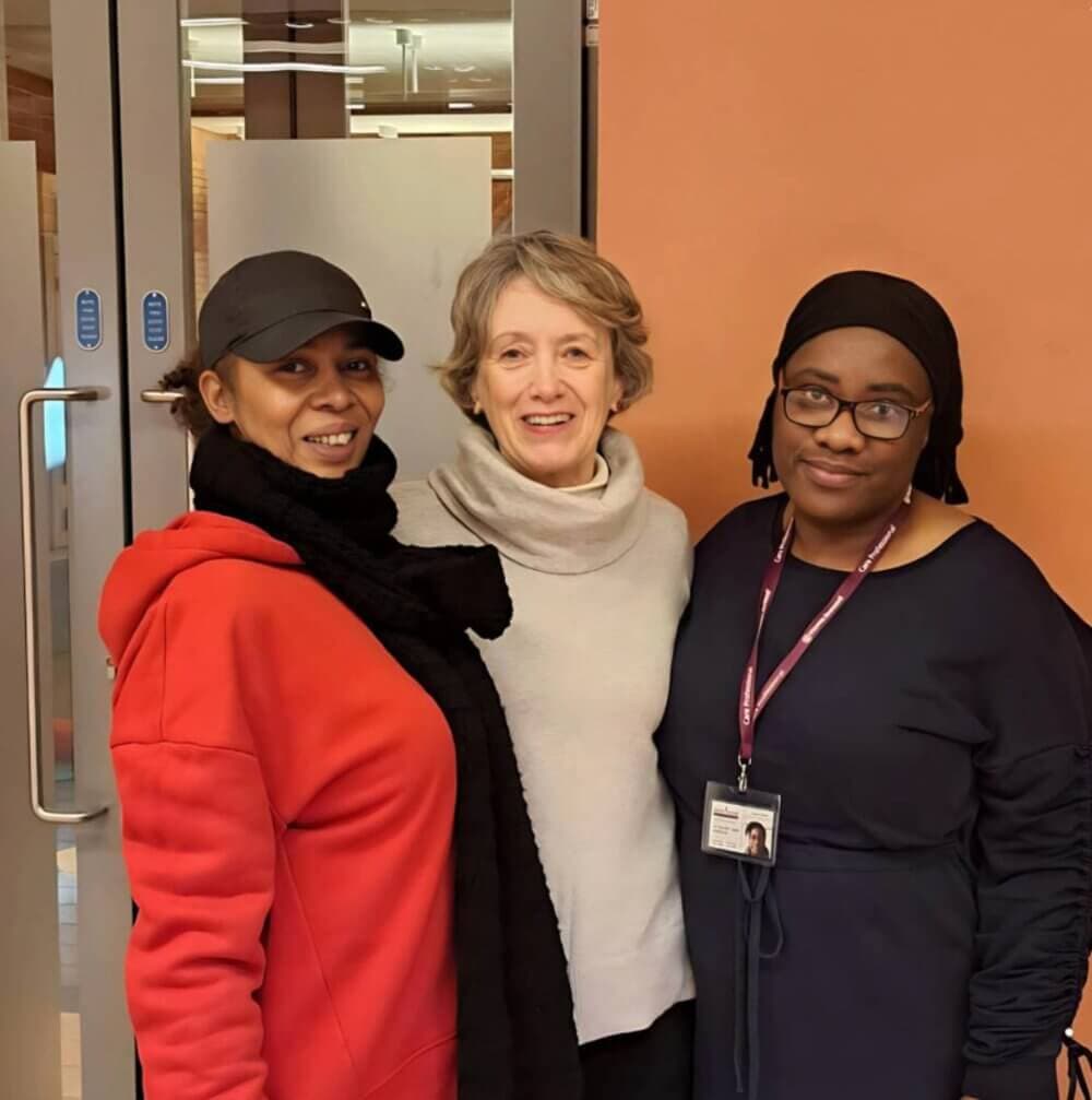 Three women stand together, two smiling, one with a neutral expression. They are indoors in front of glass doors. - Home Instead