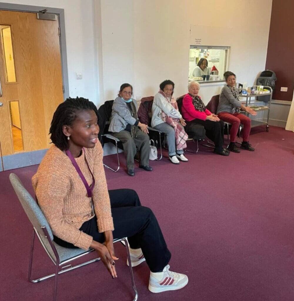 A woman is seated in front of four elderly women sitting in a line against a wall in an indoor setting. - Home Instead