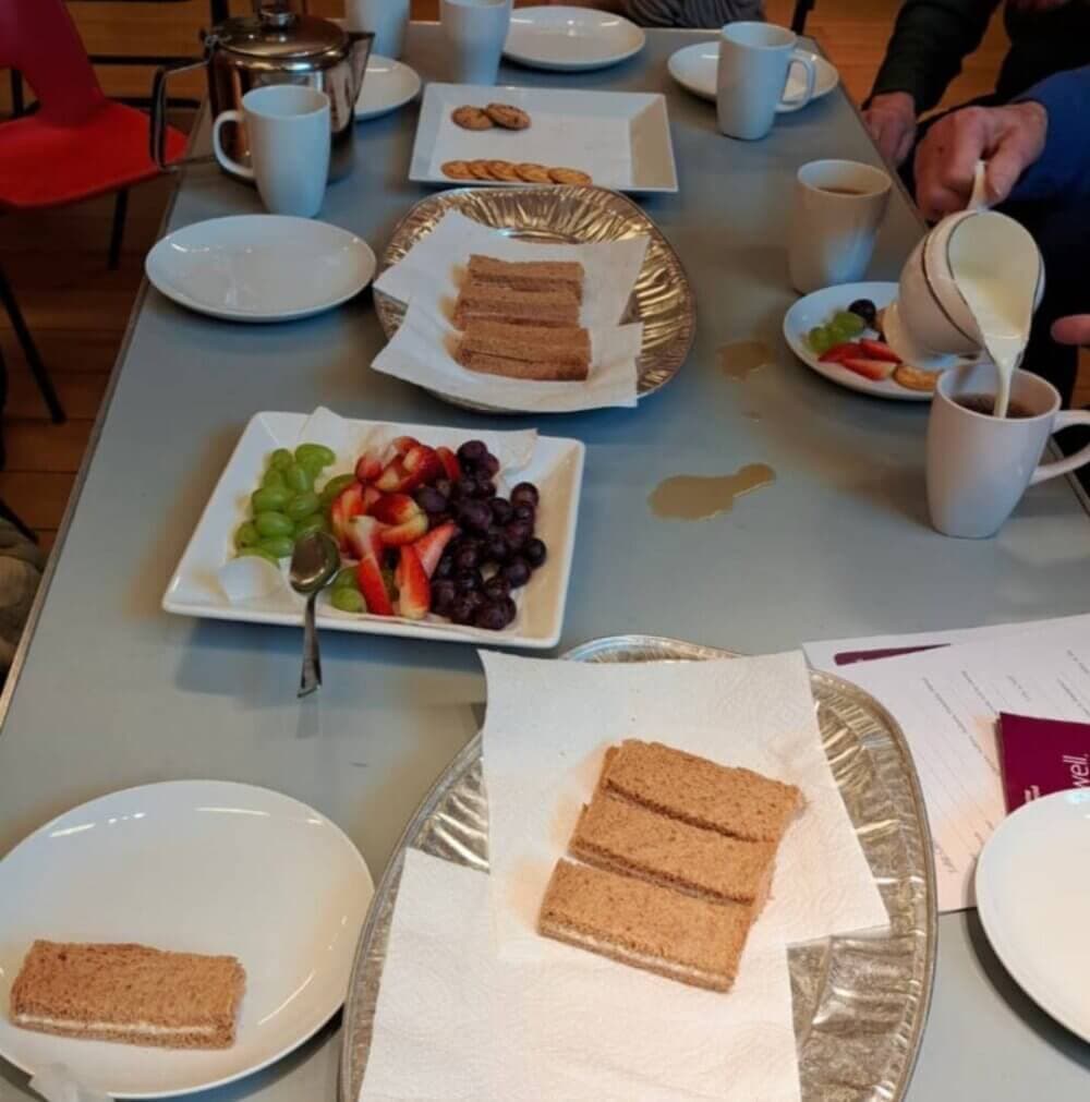 A table with plates of fruit, crackers, and bread sandwiches, with people serving coffee and tea in the background. - Home Instead