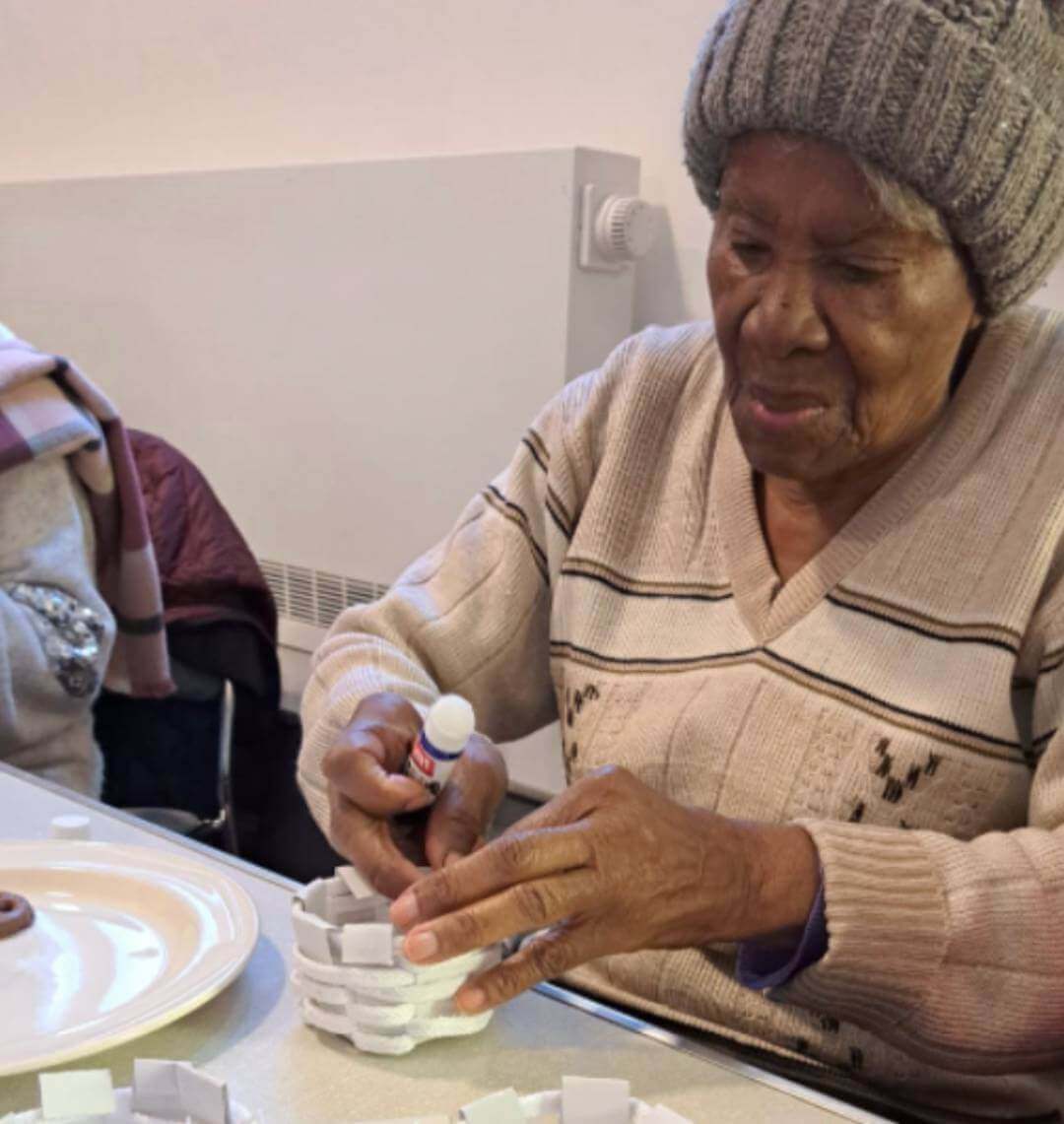 An elderly woman, wearing a gray beanie, glues pieces to a craft project at a table indoors. - Home Instead