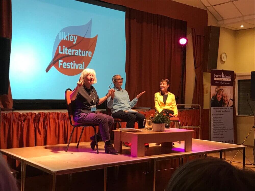 Three people seated on stage in front of an Ilkley Literature Festival screen, engaged in a lively discussion. - Home Instead