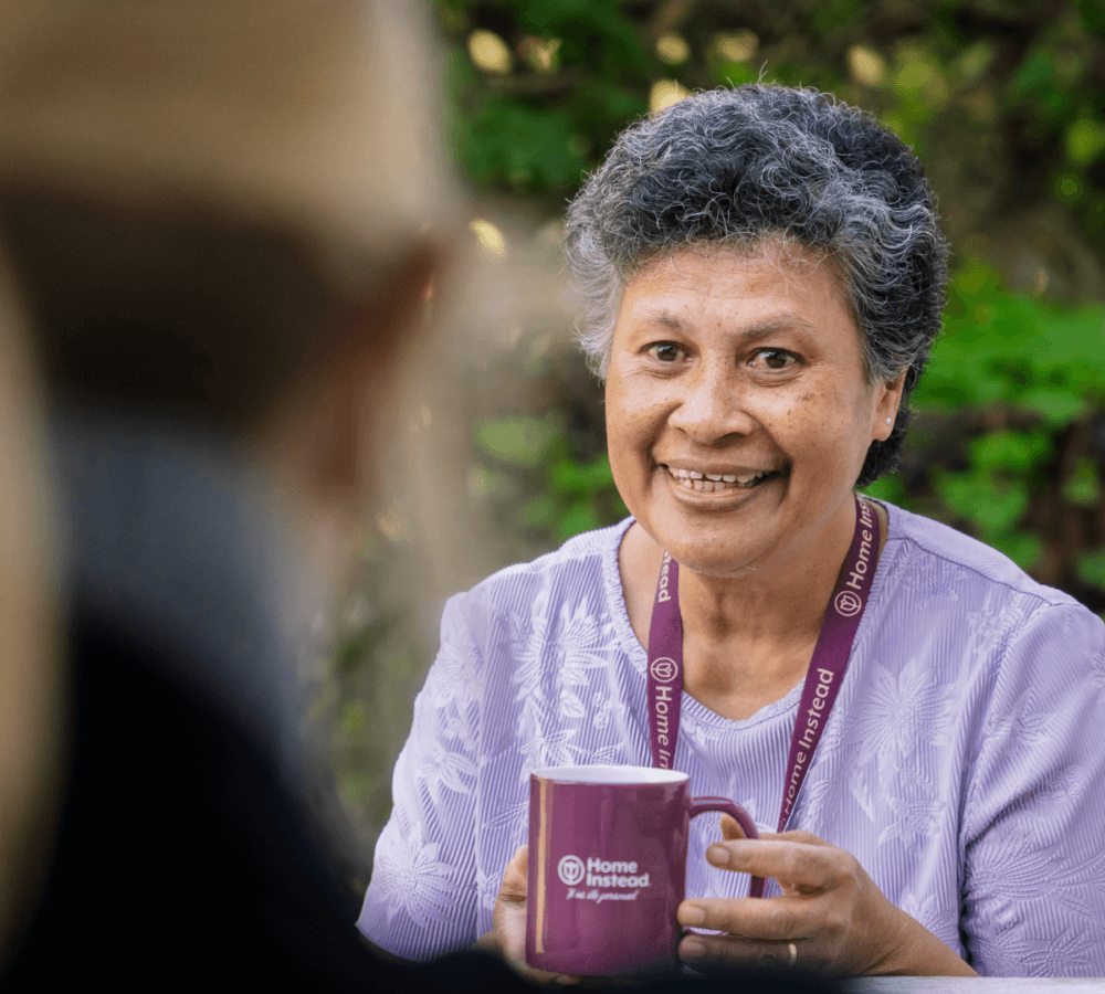 Person smiling, wearing a "Home Instead" lanyard and holding a "Home Instead" mug with a blurred figure in the foreground. - Home Instead