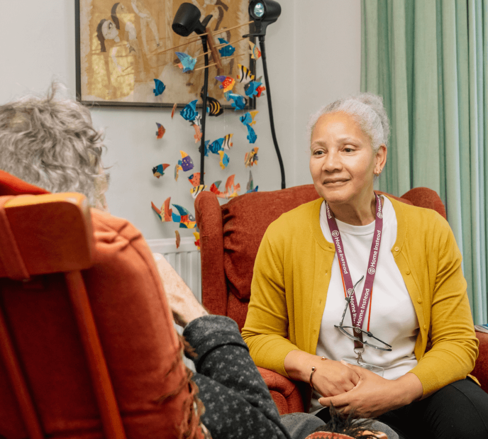 A caregiver in a yellow cardigan talks to a seated elderly person in a room with colorful decorations. - Home Instead
