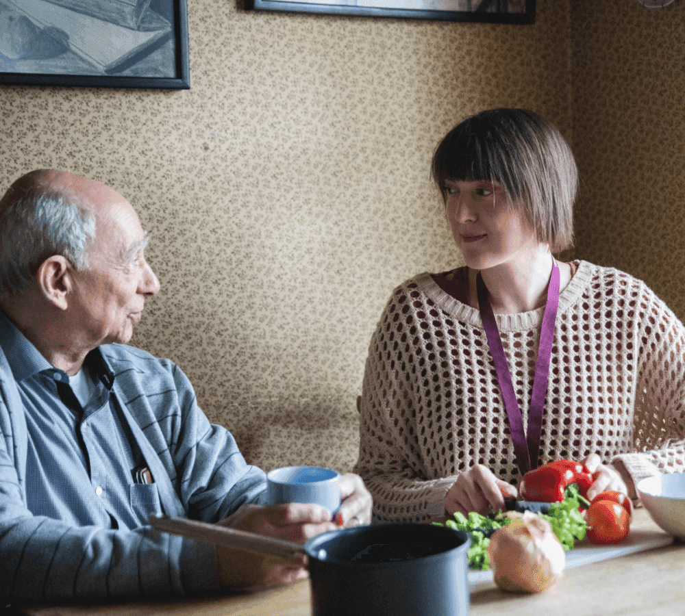 An elderly man and a young woman converse at a table with vegetables, a pot, and a cup in a cozy, wallpapered room. - Home Instead