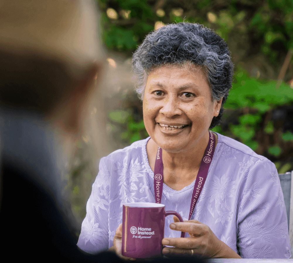 Smiling person with short, curly hair holds a purple "Home Instead" mug, wearing a matching lanyard, sitting outside. - Home Instead