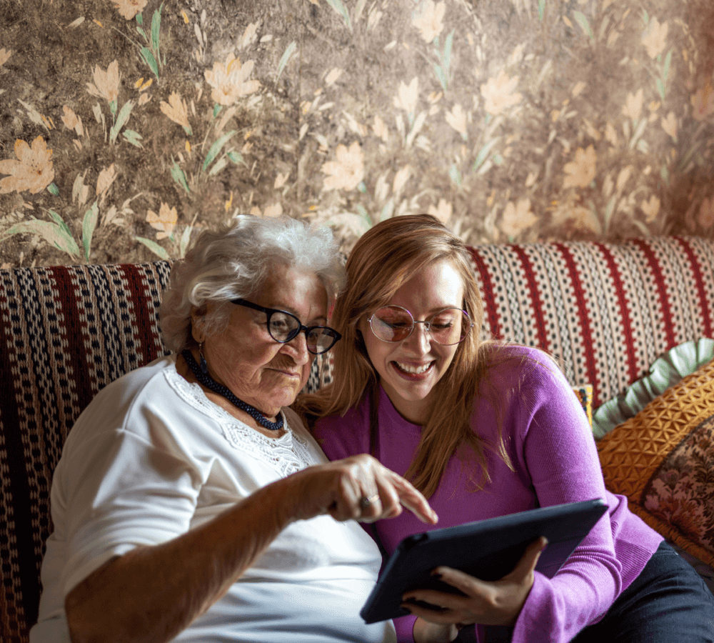 An elderly woman and a young woman sitting on a couch, smiling and looking at a tablet together. - Home Instead