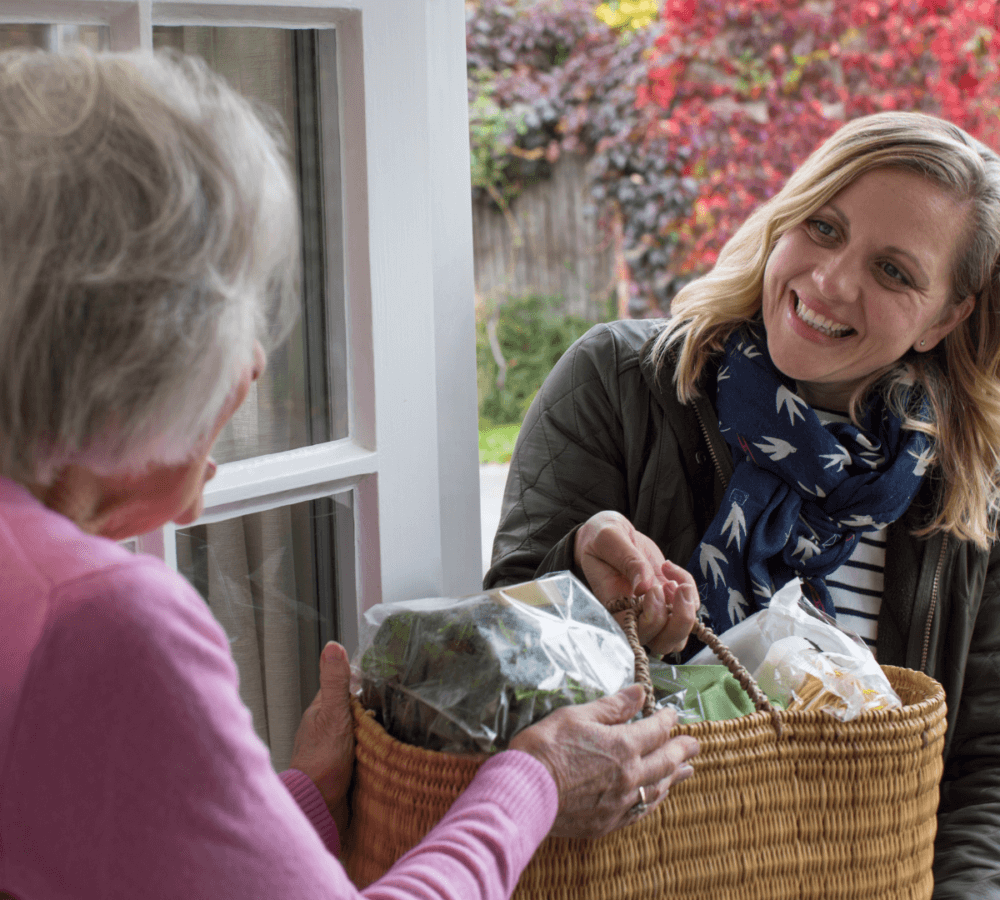 A woman delivers a basket of groceries to an elderly woman at her front door, both smiling warmly. - Home Instead