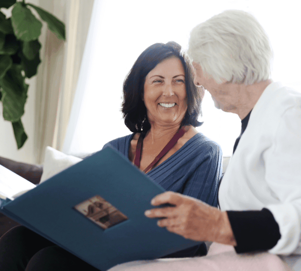 Two women smiling at each other while looking at a photo album together, sitting in a well-lit room. - Home Instead