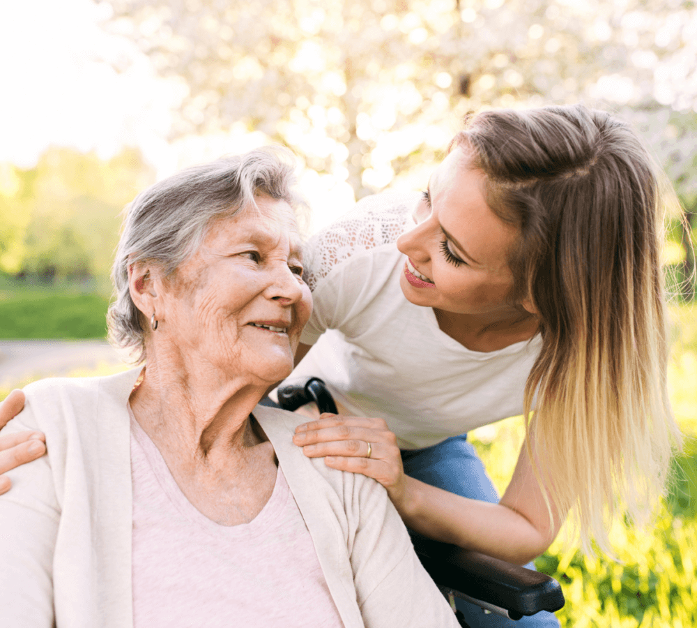 A young woman smiles and embraces an older woman in a wheelchair in a sunny, green outdoor setting. - Home Instead