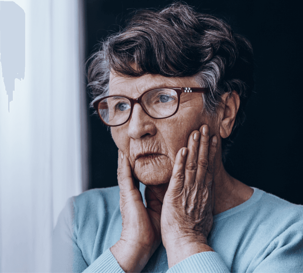 Elderly woman with glasses looking thoughtful, resting her face in her hands, wearing a blue sweater. - Home Instead