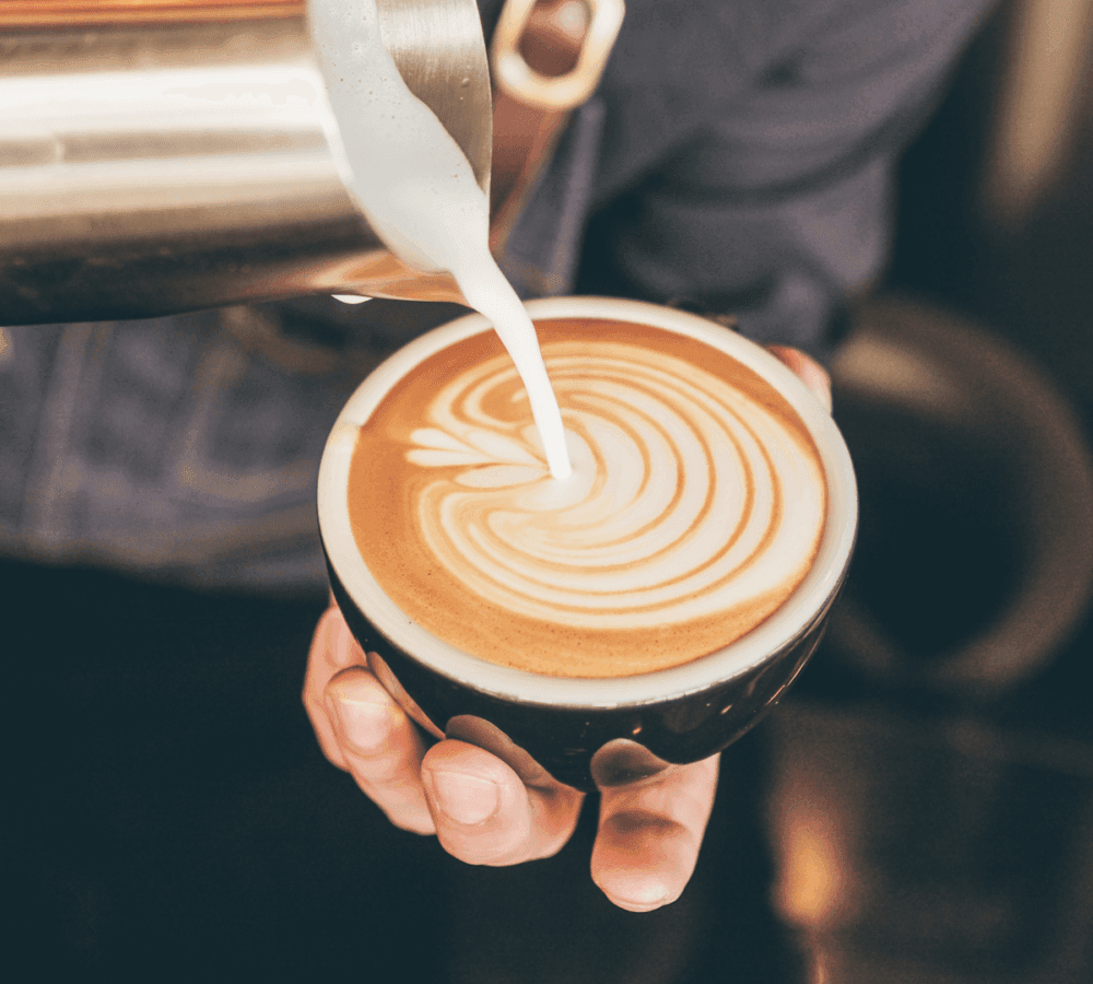 A barista pours milk into a cup of coffee, creating intricate latte art with a leaf design. - Home Instead