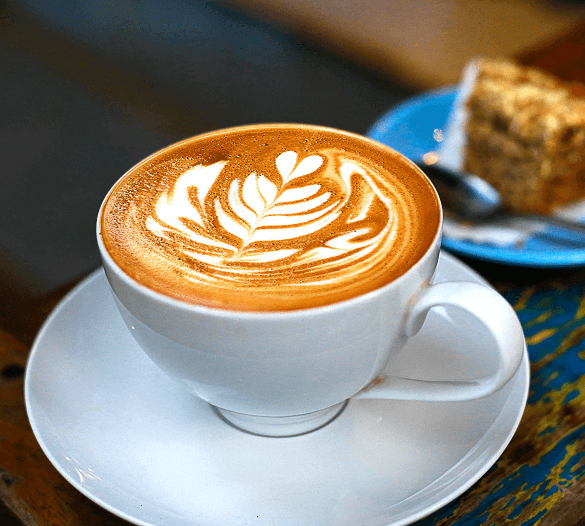 A cup of latte with intricate leaf latte art, placed on a white saucer next to a slice of cake on a blue plate. - Home Instead