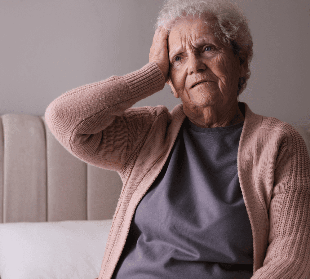 Elderly woman sitting on a bed, wearing a pink cardigan and gray shirt, with her hand on her forehead, looking worried. - Home Instead
