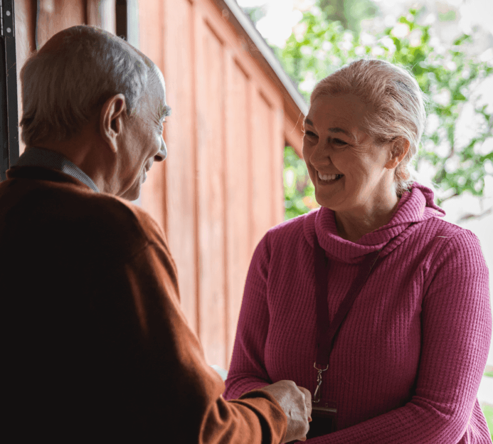An elderly man and a woman wearing a pink sweater smile and shake hands near a wooden building. - Home Instead