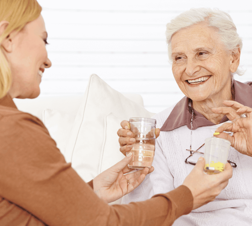 Elderly woman with glasses smiles, taking medicine and water from a younger blonde woman seated beside her. - Home Instead