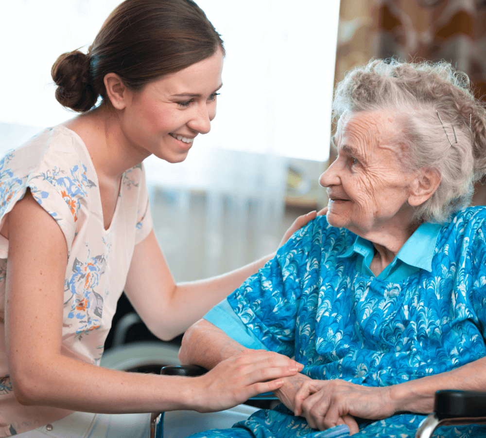 Caregiver smiling and holding hands with an elderly woman in a wheelchair, both sharing a moment of joy. - Home Instead