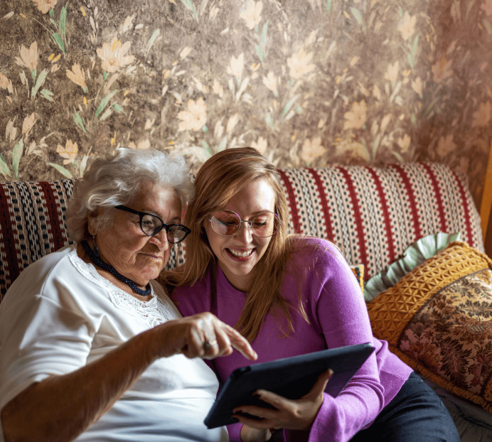 Elderly woman and young woman sit on a sofa, smiling and looking at a tablet together in a cozy, vintage-style room. - Home Instead
