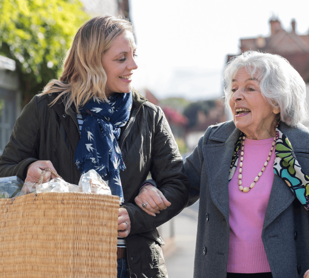 A younger woman helps an older woman carry groceries outside, both are smiling and walking arm in arm. - Home Instead