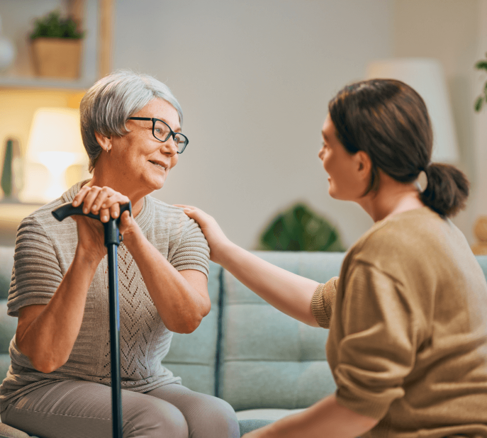 An elderly woman with a cane being comforted by a young woman with a hand on her shoulder while sitting on a couch. - Home Instead