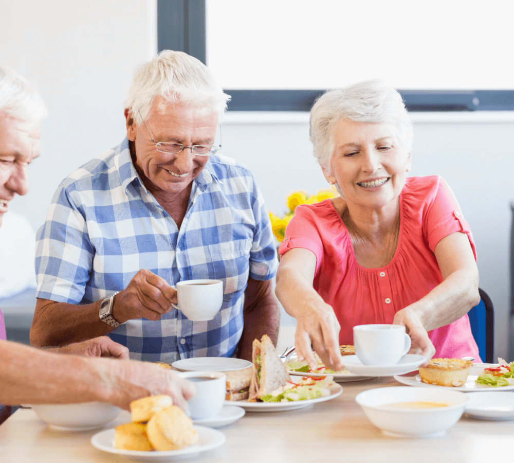Three elderly people enjoying tea and sandwiches together at a dining table. - Home Instead