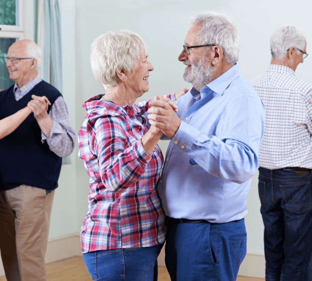 Two elderly couples are dancing and smiling in a bright room, with one couple in focus and the other in the background. - Home Instead