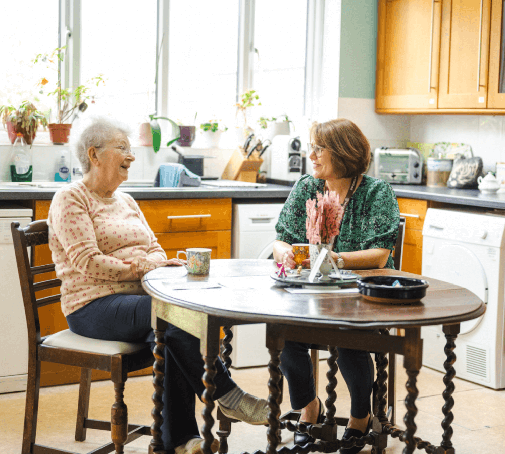 Two women seated at a kitchen table, enjoying a conversation and coffee in a brightly lit kitchen. - Home Instead