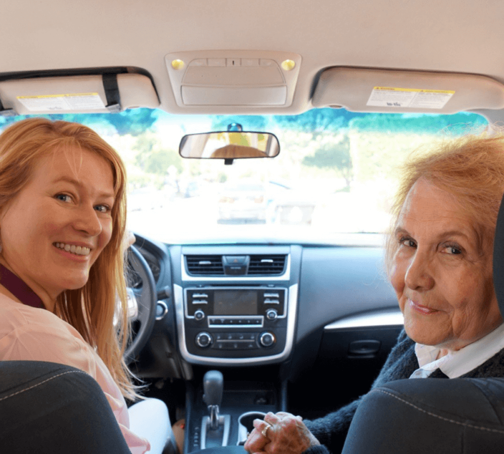 Two women sitting in a car, smiling at the camera through the front seats. - Home Instead