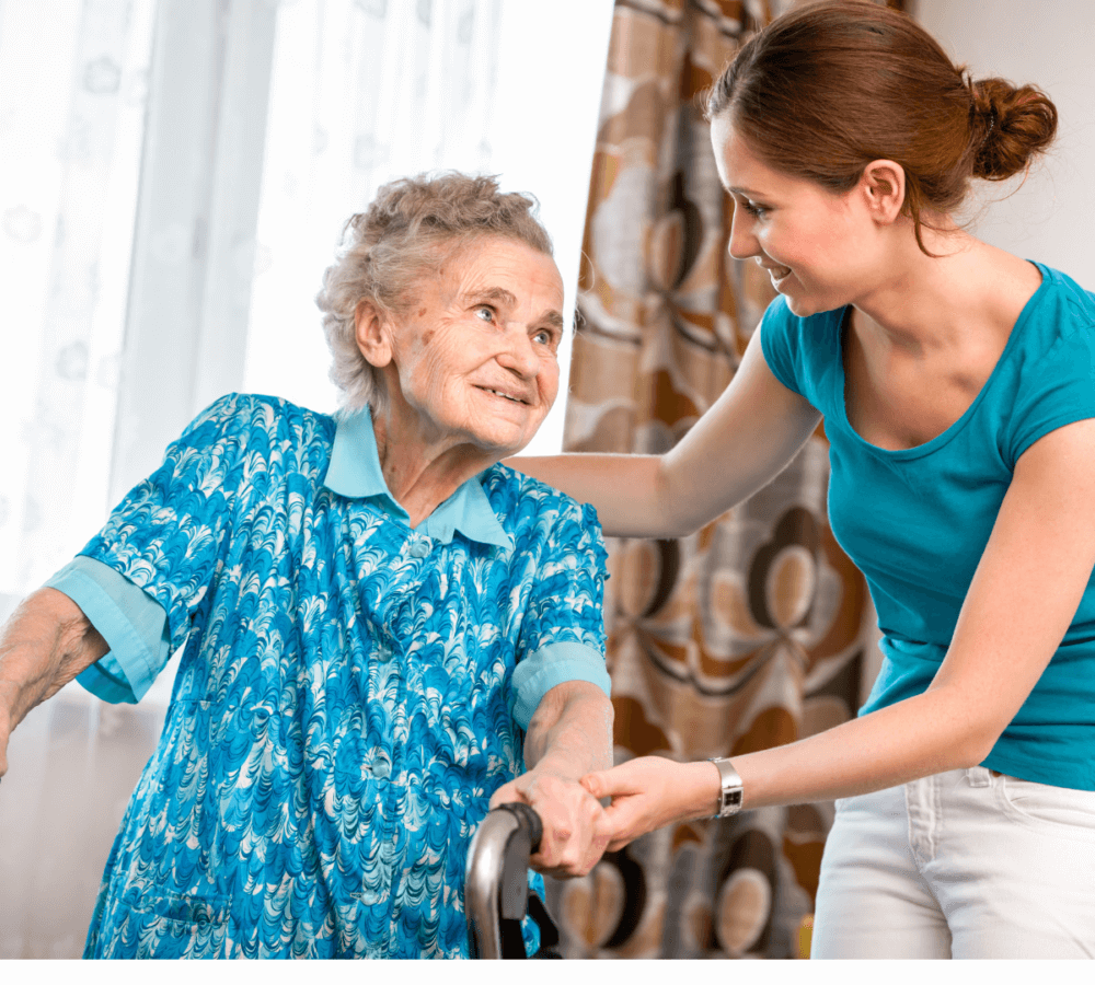 A young woman helps an elderly woman with a walker, both smiling at each other. - Home Instead
