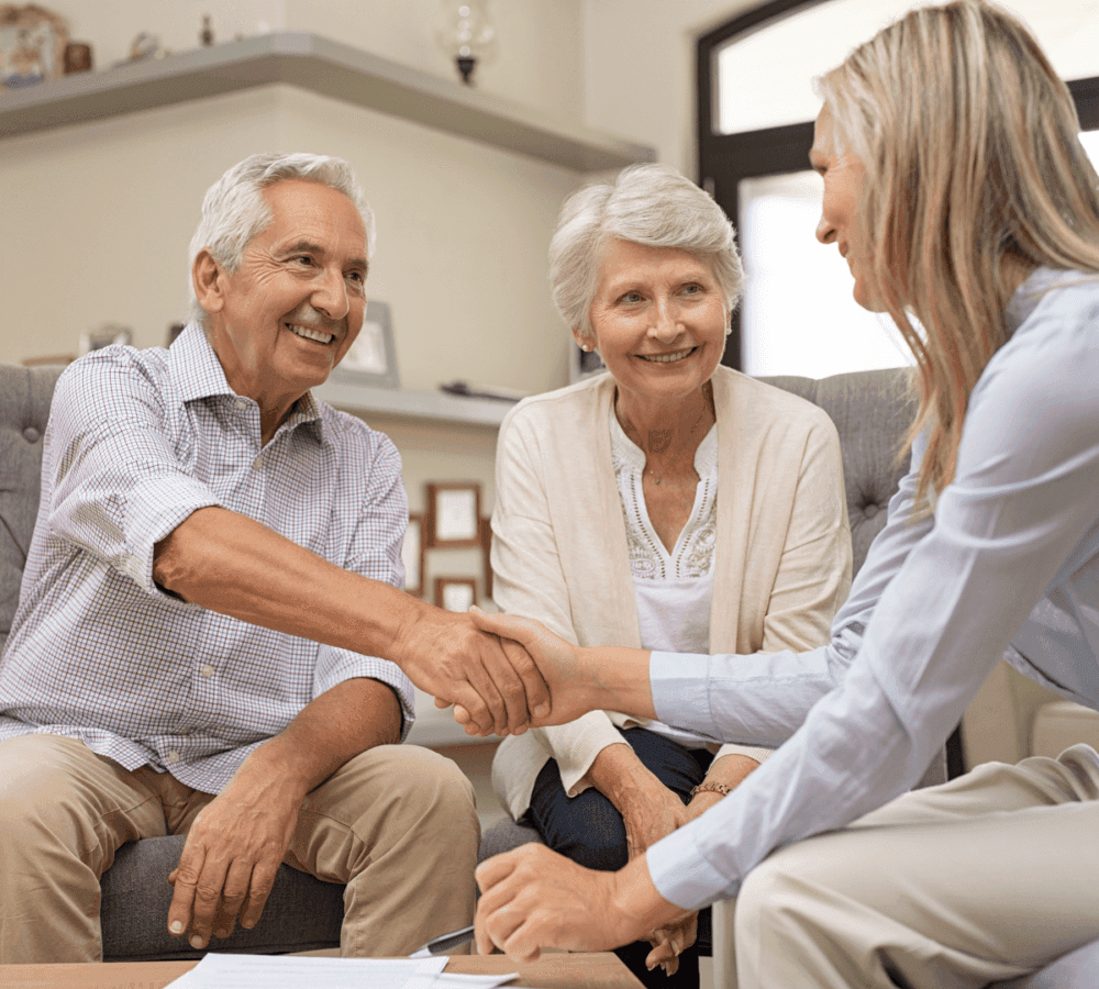 Older couple smiling and shaking hands with a woman in a living room setting. - Home Instead