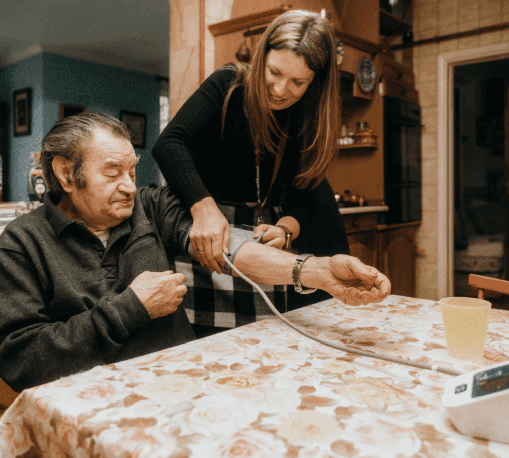 A young woman measures an older man's blood pressure at a table with a floral tablecloth in a cozy kitchen. - Home Instead