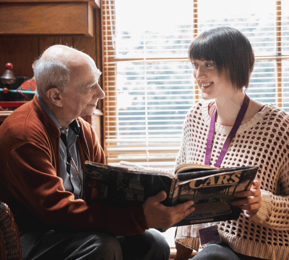 An older man and a younger woman sit by a window, smiling and reading a "CARS" magazine together. - Home Instead