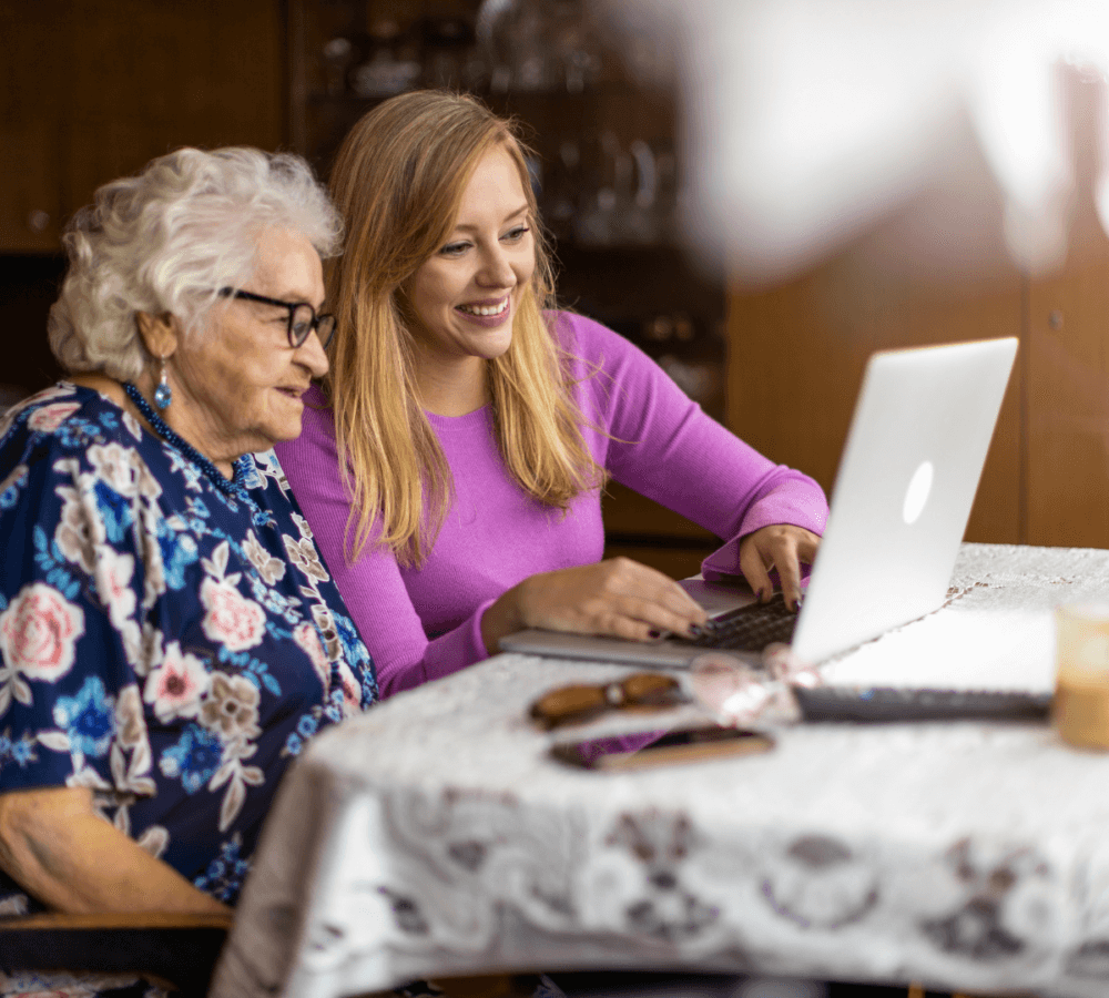 Two women, one elderly and one younger, smiling while using a laptop at a table covered with a white tablecloth. - Home Instead