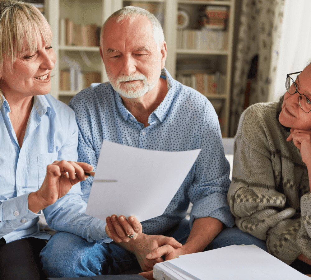 Three older adults, two women and one man, sit on a sofa looking at a document, appearing engaged in conversation. - Home Instead