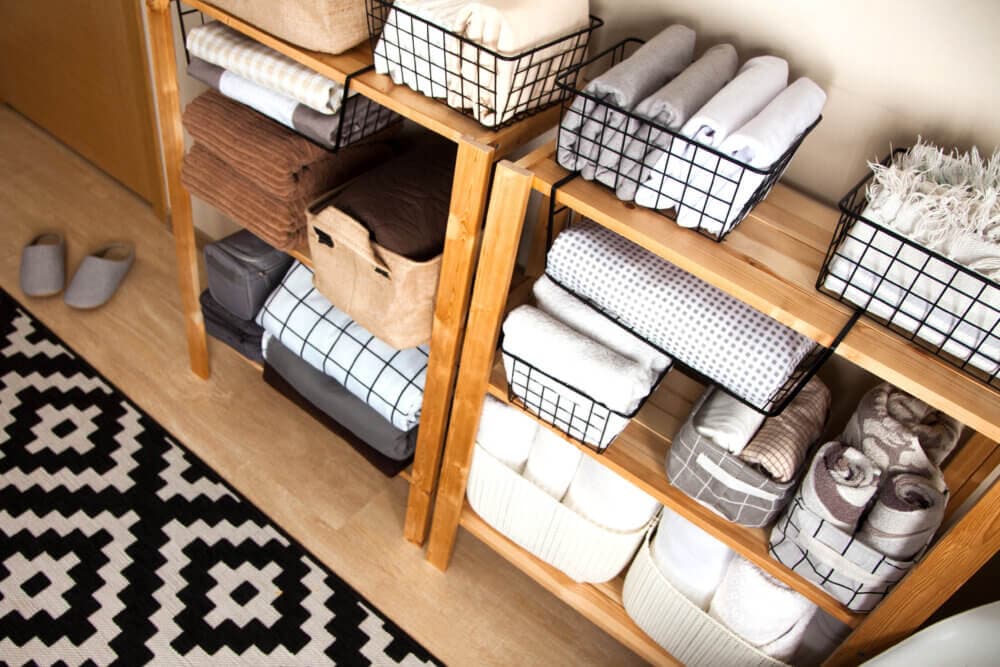 Wooden shelves with neatly organized baskets of folded towels and blankets, next to slippers and a patterned rug. - Home Instead