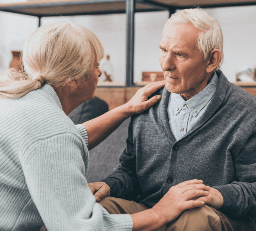 An elderly woman gently comforts an elderly man, placing her hand on his shoulder and holding his hands. - Home Instead