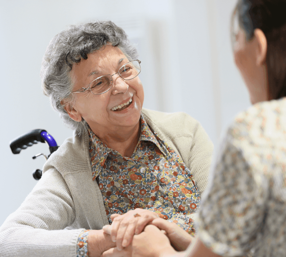 An elderly woman in a wheelchair smiles and holds hands with a caregiver, both wearing patterned shirts. - Home Instead