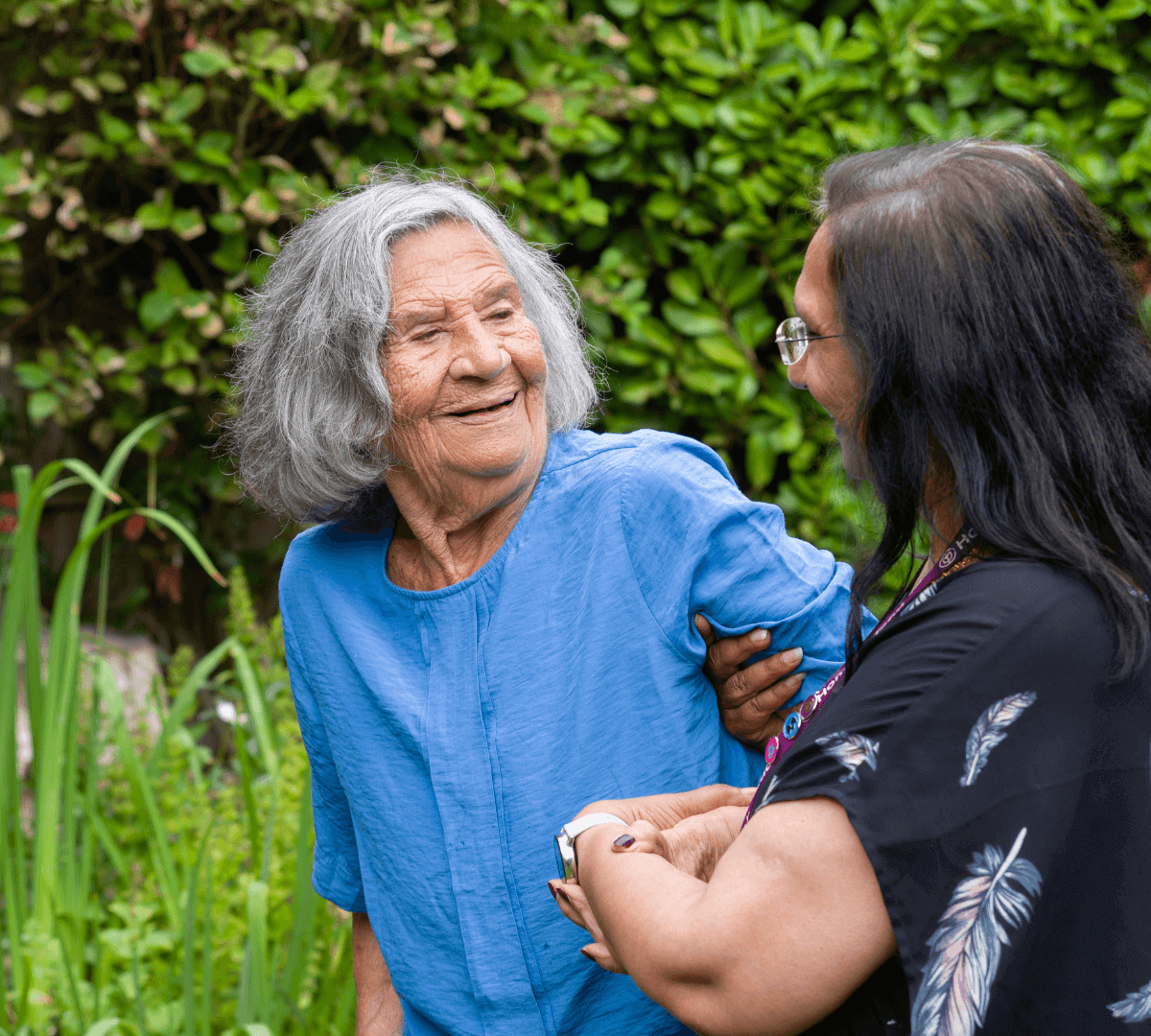 An elderly woman in a blue blouse smiles while holding hands with a younger woman in black, set against a leafy backdrop. - Home Instead
