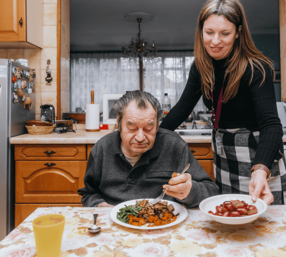 Elderly man eating at a table while a woman serves him food in a kitchen. - Home Instead