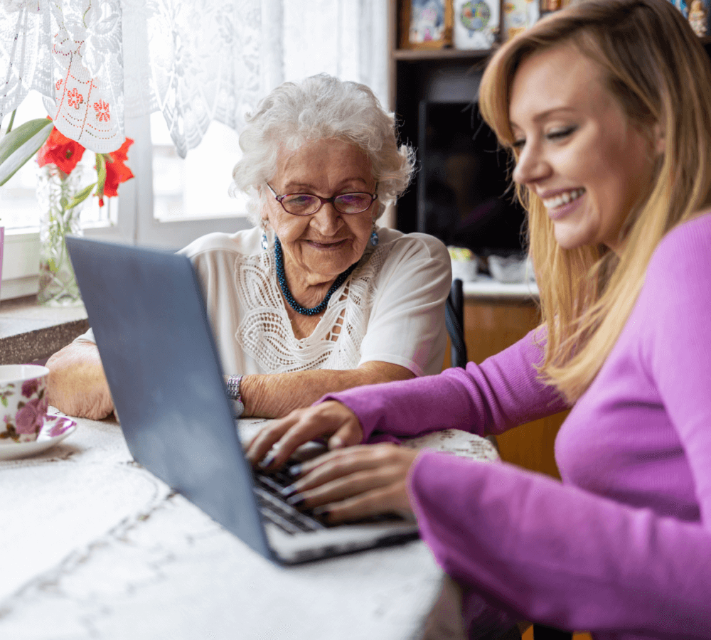 Elderly woman and young woman smiling while using a laptop together on a table near a window with lace curtains. - Home Instead