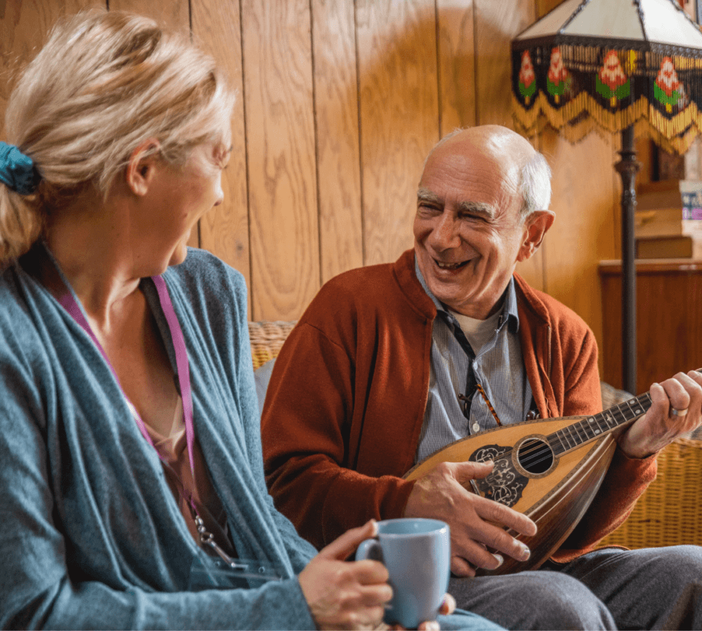 Two people sitting and smiling; an elderly man plays a mandolin, and a woman with a mug looks at him. - Home Instead