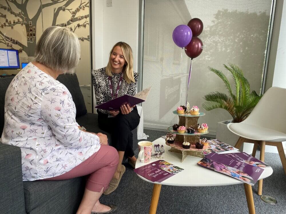 Two women sit in an office, one holding brochures. Balloons and cupcakes are on a table in front of them. - Home Instead