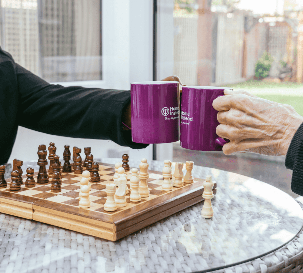 Two people clink purple mugs over a chessboard on a glass table in a sunny room. - Home Instead