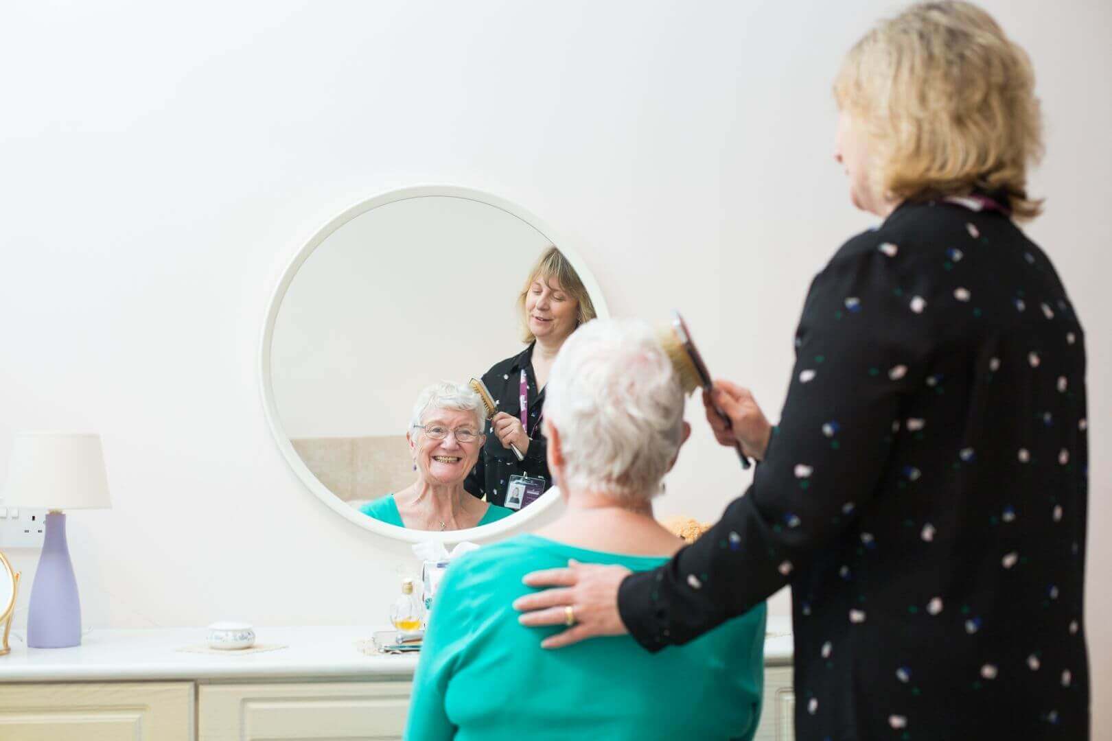 A woman combs an elderly woman's hair while they both smile at their reflection in the mirror. - Home Instead