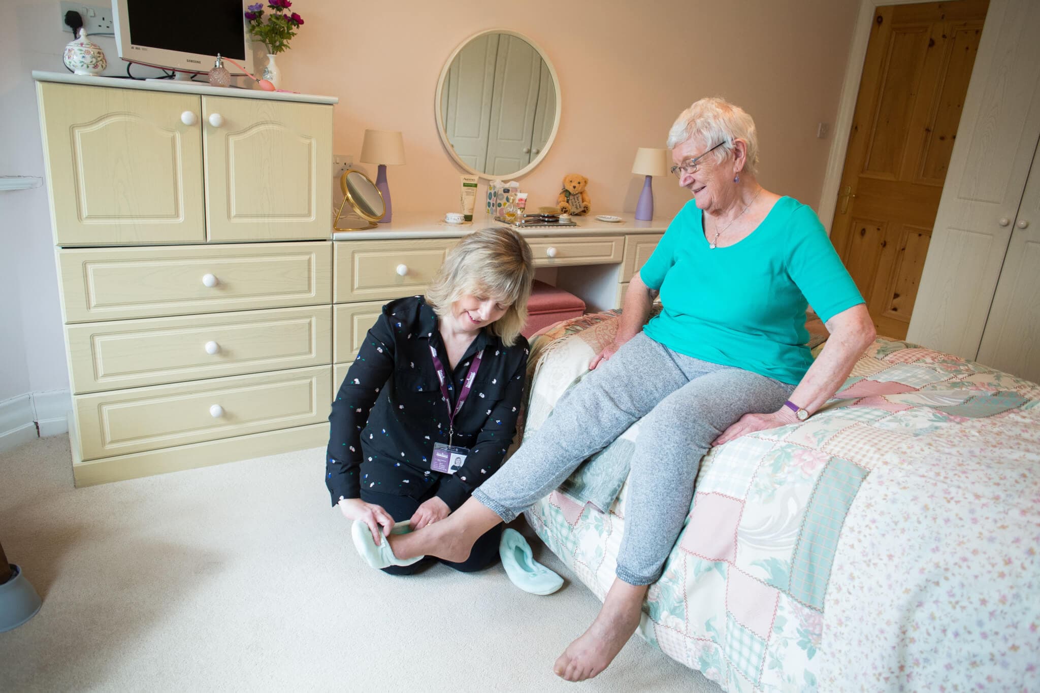 A caregiver helps an elderly woman put on slippers in a cozy bedroom. They both appear to be smiling. - Home Instead