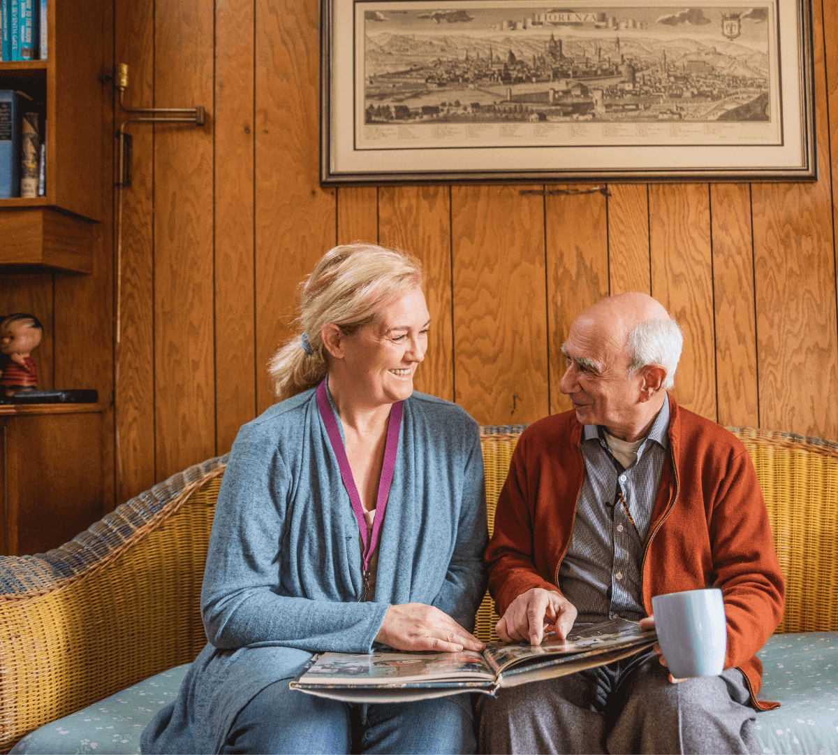 A woman and an elderly man sit on a couch, smiling and looking at a photo album together in a wood-paneled room. - Home Instead