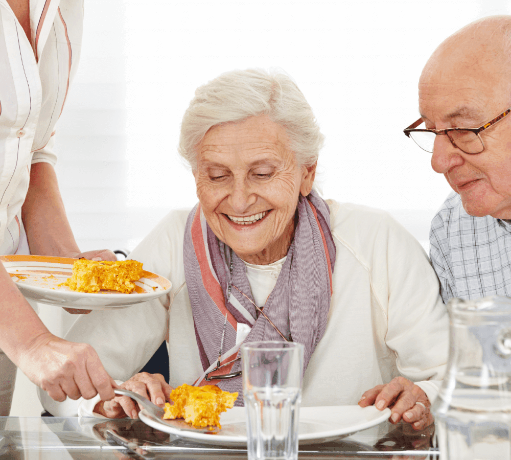Elderly woman smiling while being served food at a dining table, with an elderly man sitting next to her. - Home Instead